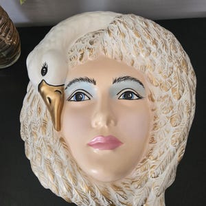 May include: A ceramic wall mask depicting a woman's face with a swan. The face has blue eyes, pink lips, and a cream complexion. The swan's beak is gold, and the hair is white with gold accents. A decorative wall hanging.