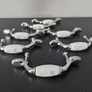 Peut inclure: Ensemble de sept poignées de meubles. Elles présentent un centre en céramique blanche orné d'un motif floral violet et vert, et des fixations en métal argenté. Chaque poignée mesure environ 10 cm de long.