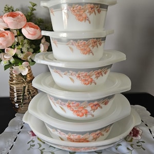 May include: A set of white serving bowls with lids, decorated with a floral pattern. Each bowl has a grey band with peach-coloured flower designs. The bowls are stacked on a white, scalloped-edge placemat, with a vase of pink flowers in the background.