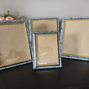 Könnte beinhalten: Ein Set aus drei Bilderrahmen mit blau-schwarzer Marmoroptik und einem dünnen goldenen Innenrand. Die Kollektion umfasst zwei quadratische Rahmen und einen rechteckigen Rahmen, die alle eine seilartige Textur am inneren Goldrand aufweisen.