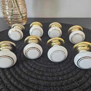 May include: Ten white and gold door knobs with a classic design. Each knob has a white, rounded body with a gold rim and a gold base. The knobs are arranged on a dark, textured surface. A decorative vase is in the background.