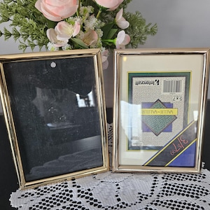 May include: Two gold-coloured rectangular picture frames. One frame is empty, the other contains a print with the text "Intercraft" and "Value-Valeur". The frames are on a white lace doily, with a floral arrangement in the background.
