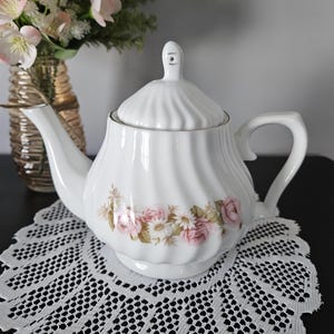 May include: White porcelain teapot with a floral rose design. The teapot has a curved spout, a handle, and a lid with a small knob. The teapot is on a white lace doily. A vase of pink flowers is in the background.