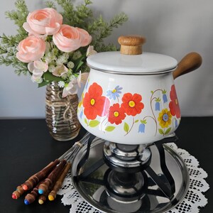 May include: A vintage white fondue pot with a floral design in red, yellow, and blue. It has a wooden handle and lid knob. The pot sits on a metal stand with a burner, and includes fondue forks. A bouquet of pink flowers is in the background.