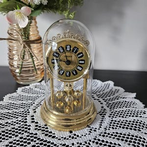 May include: A gold-coloured quartz clock with a glass dome, featuring a black and gold clock face with Roman numerals. The clock has a pendulum and is set on a gold base. A vase of flowers is in the background.