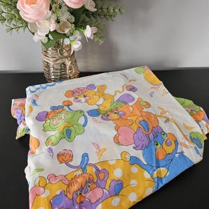 May include: A vintage white bed sheet with cartoon animal characters in blue, green, orange, and purple. The sheet has a yellow polka dot pattern and is folded. A vase of pink flowers is in the background.