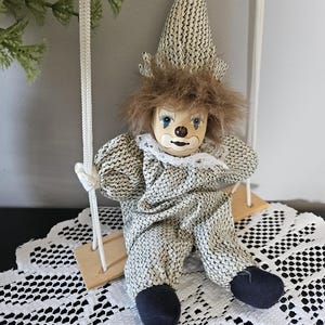 May include: A porcelain clown doll with a painted face, brown hair, and a pointed hat, wearing a textured beige and black patterned outfit with a white lace collar. The doll is seated on a small wooden swing suspended by white ropes.