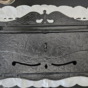 May include: A black, antique-style mailbox with a textured surface and decorative details. The mailbox features a keyhole, a decorative top, and a scalloped bottom edge. The design includes floral and scrollwork patterns.