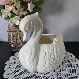 May include: A cream-coloured ceramic swan planter with detailed feather and wing patterns. The swan has a long, curved neck and a hollowed-out body for plants. A vase of pink flowers sits in the background.