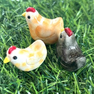 May include: Three ceramic chicken figurines, one orange, one white, and one gray, on a green artificial grass background.