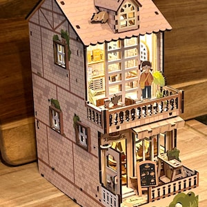 May include: A miniature wooden dollhouse with a light brown exterior and a white roof. The dollhouse has a balcony with a railing and a small figure standing on it. The dollhouse has a sign that says "Cafe" and a menu board with a list of items. The dollhouse has a small window with a light inside.