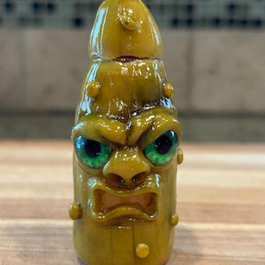 Puede incluir: Una figura artesanal en forma de pepinillo de color amarillo verdoso con una cara enfadada y ojos verdes. La figura tiene una superficie texturizada y una pequeña protuberancia curvada en la parte superior. La cara tiene la boca abierta y está rodeada de pequeños bultos redondos.