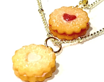 May include: Two gold chain necklaces with a cookie pendant on each. One cookie has a pink heart-shaped filling and the other has a white heart-shaped filling.