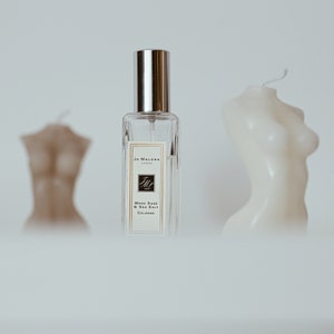 May include: A clear glass bottle of Jo Malone London cologne with a silver cap. The bottle is labeled "Wood Sage & Sea Salt Cologne". The bottle is in front of two white candles shaped like human figures.