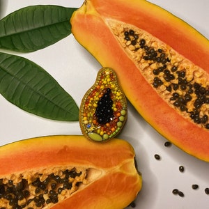 Könnte beinhalten: Eine Papaya-Brosche aus Perlen in Schwarz, Orange, Gelb und Grün. Die Brosche wird auf einer weißen Oberfläche mit zwei Hälften einer Papaya gezeigt.