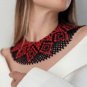 Könnte beinhalten: Eine Halskette aus Perlen mit einem geometrischen Muster in Rot und Schwarz. Die Halskette wird um den Hals getragen und hat ein detailliertes, aufwendiges Design. Der Hintergrund ist ein weißes Kleidungsstück.