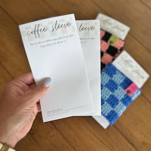 May include: A hand holds a white card with the words "coffee sleeve" and product details. The card states it fits 12-16 oz coffee cups and 12 oz cans. Crochet coffee sleeves in blue, pink, and black checkered patterns are also visible.