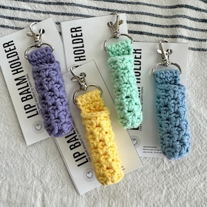 May include: Four crocheted lip balm holders in different colors: purple, yellow, green, and blue. Each holder has a silver keychain attached and a white card with the text "Lip Balm Holder" and "Fits most standard lip balm tubes."
