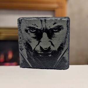 May include: A black and white image of a man's face is etched into a dark gray slate coaster. The man has a serious expression and his eyes are narrowed.
