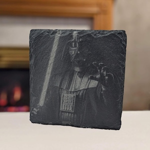 May include: A black slate coaster with a gray and white image of Darth Vader holding a lightsaber.