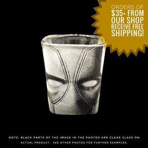 May include: A square shot glass with a Deadpool mask design. The mask is in shades of gray and black, with the eyes and facial features clearly defined. The glass is clear, and the background is black.