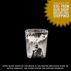 May include: A square shot glass featuring a black and white image of Batman standing in front of a cityscape. The Batman logo is visible above the city. The text on the image says "ORDERS OF $35+ FROM OUR SHOP RECEIVE FREE SHIPPING!" and "NOTE: BLACK PARTS OF THE IMAGE IN THE PHOTOS ARE CLEAR GLASS ON ACTUAL PRODUCT. SEE OTHER PHOTOS FOR FURTHER EXAMPLES."
