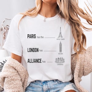 Op de afbeelding: Wit t-shirt met zwarte tekst en illustraties. Het shirt heeft de tekst "PARIS heeft de Eiffeltoren", "LONDON heeft Big Ben" en "ALLIANCE heeft Carhenge". De illustraties zijn van de Eiffeltoren, Big Ben en Carhenge.