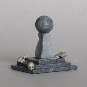 May include: A gray stone pedestal with a round top and a square base. The pedestal is decorated with skulls and bones.