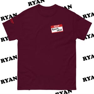 May include: A maroon short-sleeve t-shirt with the text "HELLO, RYAN'S MOM" on a name tag. The word "RYAN" is printed in black diagonally across the top and bottom of the shirt.