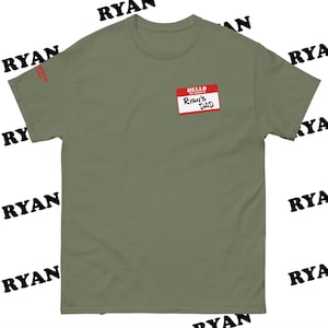 May include: Olive green t-shirt with a red and white name tag that reads "HELLO, Ryan's Dad". The shirt has a crew neck and short sleeves. The word "RYAN" is printed in black multiple times around the shirt.