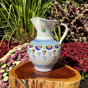 May include: A white ceramic jug with a blue and yellow floral design. The jug has a handle and a spout. It is sitting on a wooden surface.