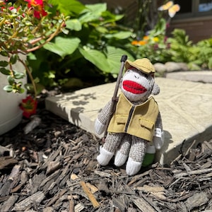 Op de afbeelding: Een sokkenaap knuffel met een beige vest en een bijpassende beige pet. De aap heeft een wit gezicht met een felrode mond, een grijs en wit gebreid lichaam en houdt een dunne bruine houten wandelstok vast. Een klein wit label is zichtbaar.