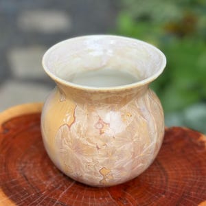 Puede incluir: Un jarrón de cerámica con cuerpo redondo y una abertura ancha. El jarrón está decorado con un patrón blanco y dorado.