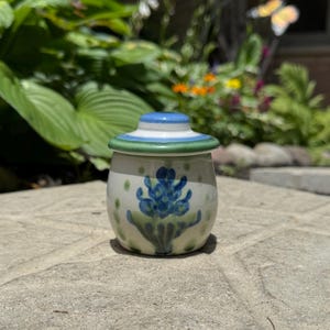 Puede incluir: Un pequeño tarro de cerámica redondo con tapa a juego, que presenta un diseño floral azul pintado a mano sobre un fondo color crema. La tapa tiene un borde azul y verde, y el tarro está decorado con pequeños puntos verdes alrededor.