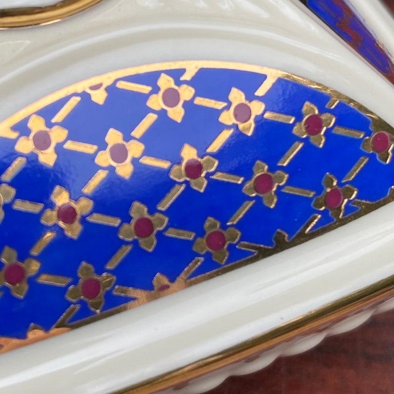 Op de afbeelding: Een close-up van een blauwe en gouden keramische sieradendoos met een decoratief patroon van kleine bloemen. De doos heeft een witte achtergrond en een gouden rand.