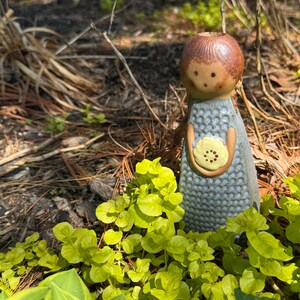 Może przedstawiać: Ceramiczna figurka w kształcie postaci z brązową głową i jasnoniebieską, teksturowaną sukienką. Postać trzyma w dłoniach mały, bladożółty, okrągły przedmiot z kropkowanym środkiem. Figurka ma prostą twarz z dwojgiem oczu.