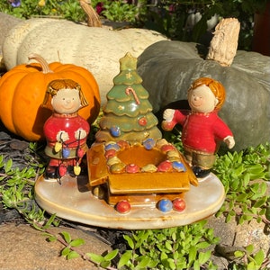 May include: A ceramic Christmas decoration featuring a Christmas tree, two children, and a box of ornaments. The tree is green with a star on top. The children are wearing red sweaters. The base is a light brown color.