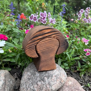 May include: A wooden mushroom-shaped box with a dark brown finish, resting on a gray rock. The mushroom has a textured, layered design. The background features a vibrant garden with colorful flowers.