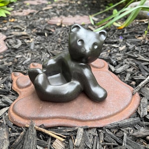 May include: A dark gray bear figurine sitting on a brown, foot-shaped base. The bear is in a seated position with its arms and legs curled. The base has a textured surface, and the bear has visible facial features.