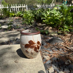 May include: A ceramic vessel with a rounded body and a tapered base, featuring a speckled off-white glaze. The rim is finished in a dark brown tone, and a hand-painted brown botanical motif of leaves on a stem wraps around the exterior surface.