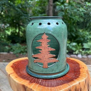 Puede incluir: Un portavelas de cerámica verde con un diseño de árbol recortado. El soporte tiene una base redonda y un cuerpo cilíndrico con varios agujeros pequeños cerca de la parte superior. El árbol es de color marrón rojizo y está centrado en la parte delantera. El portavelas está sobre una superficie de madera.