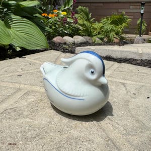 May include: A white ceramic duck figurine featuring a smooth, glossy finish with delicate blue painted accents along the head, back, and wings. The bird is depicted in a resting position with a rounded body, a small beak, and a distinct crest.