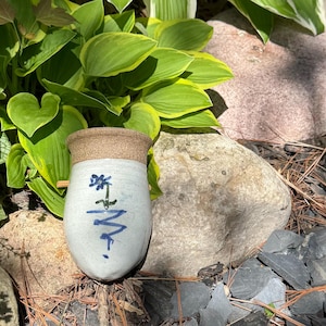 Può includere: Un vaso da parete in ceramica con bordo beige maculato e corpo bianco. La parte anteriore presenta un disegno di un fiore blu dipinto a mano con uno stelo verde e un motivo a zigzag blu sottostante. Un piccolo tassello in legno sporge dal lato.