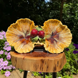 May include: A double serving dish made of ceramic with a yellow and brown glaze. The dish is shaped like two large flower petals with a raised center section featuring three red cherries with green leaves.