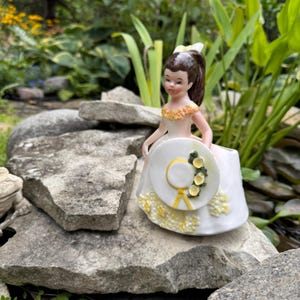 以下が含まれることがあります： 白いドレスに黄色の花のアクセント、お揃いの帽子を持った若い女性のヴィンテージ磁器フィギュア。フィギュアは茶色の髪で、自然な屋外の背景に設定されています。