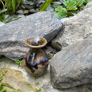 Könnte beinhalten: Eine kleine, handgefertigte Keramikvase mit breitem, ausgestelltem Rand. Die Vase hat eine glänzende Oberfläche mit einer Mischung aus Braun-, Blau- und Goldtönen. Die Vase steht auf einer Steinoberfläche, umgeben von großen Felsen und Grün.