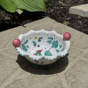 May include: A white ceramic bowl with a scalloped rim and two pink spherical handles. The interior features hand-painted teal floral motifs, small yellow and purple dots, and a central rooster illustration with a red comb and green tail feathers.