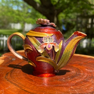 May include: A small, ornate teapot with a red, gold, and purple finish. The teapot is decorated with a flower and leaves, and has a shiny, metallic finish.