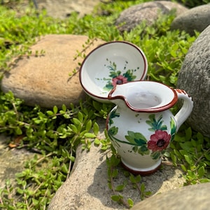 Könnte beinhalten: Ein weißes Keramik-Krug- und Schüssel-Set mit handgemalten Blumendesigns. Der Krug hat einen Griff und einen Ausguss, und beide Teile weisen rote Blumen und grüne Blätter auf. Die Ränder sind braun eingefasst.