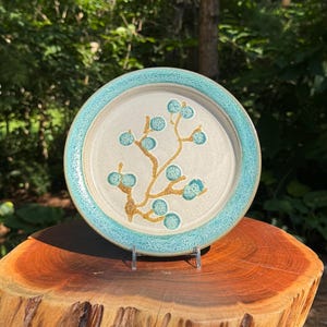 May include: A ceramic plate with a light beige center and a turquoise rim. The plate features a brown branch design with turquoise berry-like shapes. The plate is displayed on a clear stand, showcasing its decorative design.